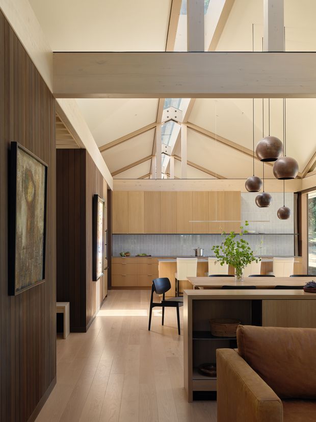 Warm, contemporary open-plan kitchen and dining area viewed from the living space. The room features extensive light timber cabinetry and flooring, dark vertical timber wall panelling, a grey tiled backsplash, exposed pale timber structural beams, clerestory glazing, and a linear skylight. A timber dining table, upholstered chairs, sculptural dark pendant lights, and a partially visible brown sofa create a refined, natural interior.