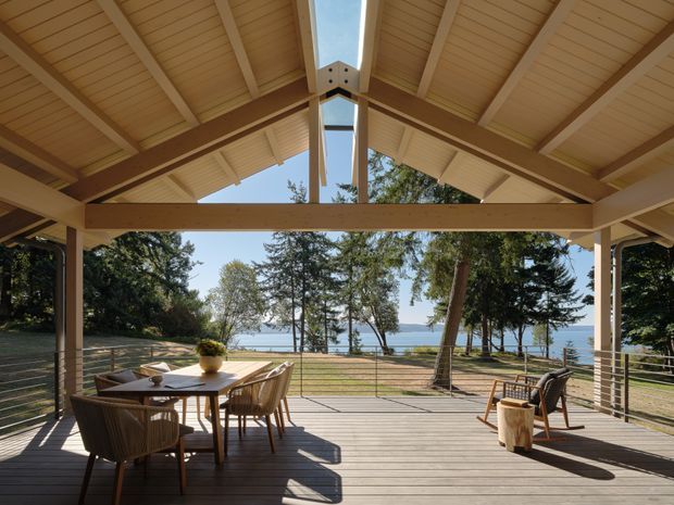 A spacious covered outdoor entertaining pavilion with a pitched timber roof, exposed timber beams and rafters, and a central linear skylight. The pavilion opens onto a timber deck overlooking a landscaped waterfront setting, with a dining table and chairs on the left and lounge seating on the right. The open structure creates a bright, relaxed indoor-outdoor living space.