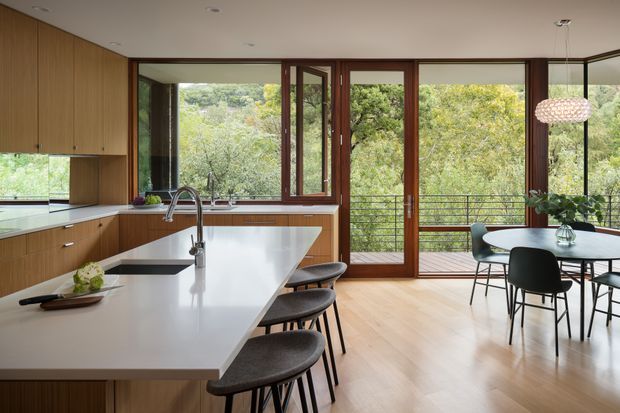 A bright open-plan kitchen and dining area with extensive warm timber cabinetry, a large white island with integrated sink and seating, and a round dining table beside full-height glazed timber doors opening to a balcony. The room overlooks leafy greenery and features a contemporary pendant light, dark-framed dining chairs, and pale timber flooring.