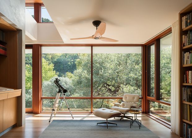 A bright, refined sitting and reading room with expansive floor-to-ceiling timber-framed glazing overlooking dense greenery. Built-in timber bookshelves line both sides, while a mid-century lounge chair and ottoman sit on a large rug beside a telescope. A ceiling fan, warm timber flooring, and a clerestory opening enhance the airy, indoor-outdoor feel.