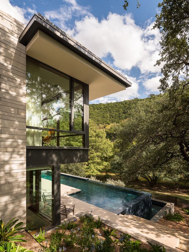 Exterior architectural photograph of a contemporary multi-level home set among dense wooded hills. The image focuses on a projecting upper-level volume with extensive dark-framed glazing, pale horizontal wall cladding, and a broad flat roof overhang. A swimming pool and paved terrace occupy the foreground, surrounded by mature trees and landscaped planting.
