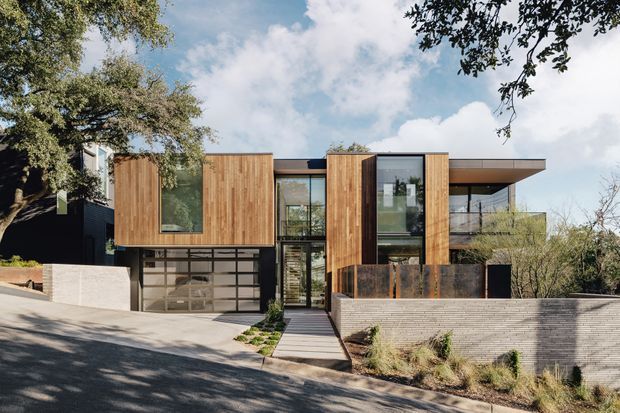 Street-facing exterior view of a contemporary multi-level residence on a sloped site. The facade combines warm vertical timber cladding, extensive floor-to-ceiling glazing, dark metal framing, a projecting flat roof, and a glazed garage door. A paved path leads from the street to the entry, with landscaped planting and a low light-brick retaining wall in the foreground.