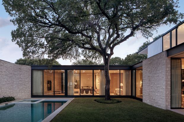 A contemporary single-storey courtyard residence arranged around a rectangular swimming pool and mature oak tree. The home combines pale textured masonry walls with dark metal-framed floor-to-ceiling glazing, creating a strong connection between the interior living spaces and landscaped courtyard. Warm interior lighting is visible through the glass, while a raised, angular roof volume appears at the right.