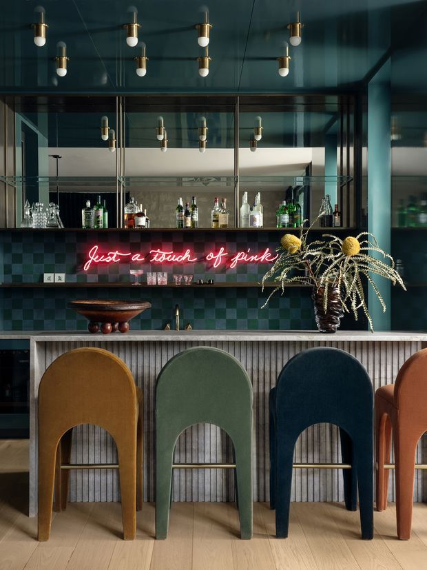 A sophisticated home bar with a dark teal ceiling and walls, mirrored open shelving displaying liquor bottles and glassware, and a deep green-blue tiled backsplash. A pale stone or composite counter sits above vertically ribbed bar cabinetry, with a row of sculptural upholstered bar stools in ochre, sage green, navy, and terracotta. Brass pendant bulbs, a neon pink sign, and a large decorative arrangement add character and contrast. The room has a moody, richly layered hospitality-inspired atmosphere.