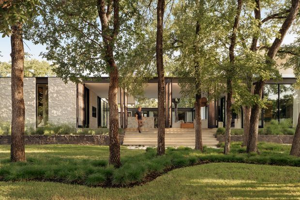 A low, contemporary pavilion-style residence viewed from a landscaped garden. The facade combines pale masonry walls, extensive dark-framed glazing, timber-toned vertical screens, and a broad covered entry terrace reached by shallow steps. Mature trees partially screen the building, with lawn and layered planting in the foreground.