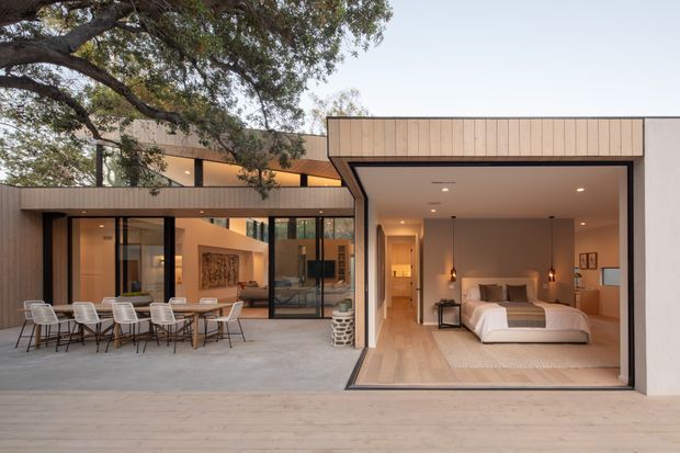 A contemporary single-storey residence organized around a broad paved outdoor terrace and a glazed courtyard-like connection. The open-fronted wing on the right contains a bedroom with a large bed, warm timber flooring, a fireplace, bedside lighting, and built-in cabinetry. Floor-to-ceiling glazing connects the bedroom and adjacent interior spaces to the terrace, while vertical timber cladding and a covered outdoor dining area sit beneath mature trees. The palette is warm and restrained, with strong indoor-outdoor connection and clean rectilinear forms.