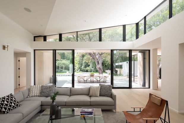 Bright contemporary open-plan living room with a large sectional sofa, glass coffee table, leather lounge chair, and extensive black-framed glazing. The room has a high ceiling with clerestory-style windows and opens directly to an outdoor dining terrace and landscaped garden. Neutral furnishings and pale timber flooring create an airy, refined interior.