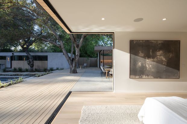 A contemporary bedroom opening directly onto a spacious timber deck and landscaped courtyard through full-height corner glazing. The room has pale timber flooring, white walls, recessed ceiling lights, a large abstract artwork, and a partially visible white bed. Outdoor paving, boundary fencing, mature trees, and a minimalist pool or water feature are visible beyond the open connection.