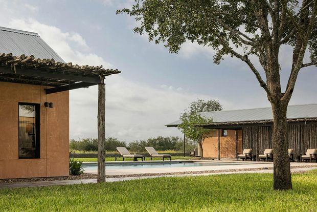 A contemporary rural-style property composed of low, single-storey buildings around a long swimming pool. The buildings feature warm earthen-toned walls, dark vertical timber screening, corrugated metal pitched roofs, and rustic timber posts supporting covered verandas. Mature trees and open landscaped lawn surround the pool, with lounge chairs and outdoor seating creating a relaxed resort-like setting.