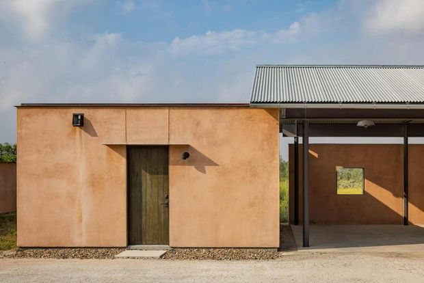 A minimalist single-storey exterior with a simple ochre rendered masonry volume and dark timber entry door, connected to a covered open-sided structure. The structure has a shallow corrugated metal roof supported by slender dark posts, with a small square window and gravel landscaping in front. The composition is clean and utilitarian, with strong geometric forms and warm earthy tones.