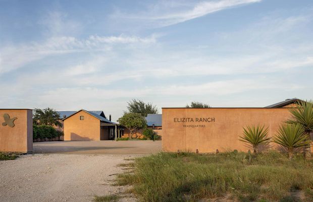Exterior view of the Elizita Ranch headquarters entrance, with a warm terracotta stucco boundary wall bearing signage, a gravel driveway, low ranch-style buildings, and drought-tolerant desert planting under a bright sky.
