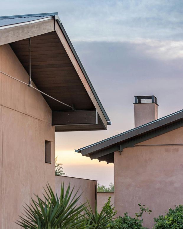 Exterior architectural detail of a low-rise home showing two intersecting stucco volumes with sloped metal roofs, broad timber-lined eaves, a chimney, and a small enclosed courtyard or passage. The warm pink-beige walls and dark roof trims are set against an overcast dusk sky with foreground planting.