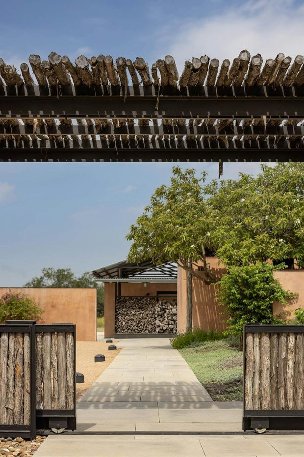 A landscaped residential entrance viewed from an open timber-and-metal gate. A straight concrete path leads toward a covered carport or storage structure filled with stacked firewood. The property uses earthy rendered walls, dark steel framing, and prominent rough timber posts, including a pergola-like overhead structure with exposed cut logs. Mature trees and gravel planting beds surround the approach under a clear blue sky.