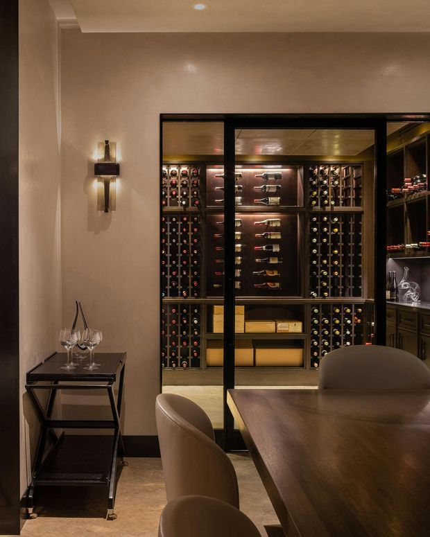 A moody, refined wine room and dining area viewed from inside. A dark timber dining table with rounded upholstered chairs occupies the foreground, while black-framed glazed doors reveal extensive built-in wine storage, bottle racks, display shelving, and illuminated bottle shelving beyond. Warm concealed lighting, taupe walls, dark joinery, and a small drinks trolley create a luxurious hospitality-inspired atmosphere.