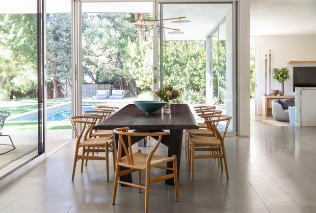 A bright, open-plan dining area centered on a dark rectangular dining table with timber armchairs. Floor-to-ceiling glazed sliding doors open the room to a landscaped garden and swimming pool, while a contemporary living area is visible beyond. The space has pale tiled flooring, white walls, abundant natural light, and a slim sculptural pendant light.