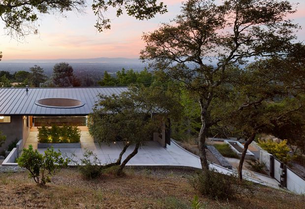 Exterior view of a modern, low-profile house integrated into a wooded, sloping site. The home features a broad metal roof with a circular skylight, expansive glazing, pale concrete terraces and retaining walls, and landscaped courtyards. Mature trees frame views across a distant valley or urban landscape at sunset.