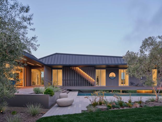 Single-storey contemporary residence viewed from the landscaped front garden, with a dark standing-seam metal roof and dark vertical cladding. Warmly lit glazed rooms open onto a paved terrace beside a rectangular swimming pool, with sculptural seating, raised planters, ornamental grasses and established trees creating a refined outdoor setting.