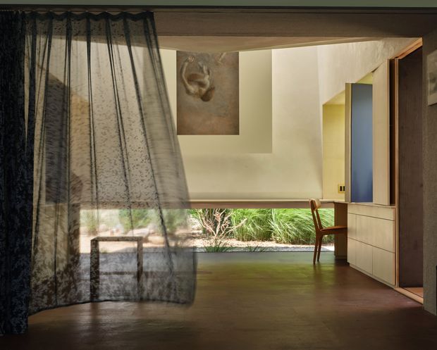 A warmly lit, low-profile interior with dark timber flooring, a broad opening to a planted outdoor area, and a minimalist built-in timber cabinet with a blue accent panel. Sheer patterned curtains frame the opening, while a simple wooden chair, recessed ceiling, artwork, and timber-framed doorway create a refined, tranquil atmosphere.
