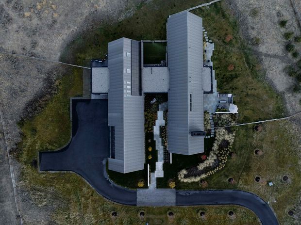 Aerial top-down view of a large contemporary building complex with two long, parallel grey metal-clad pitched roof forms connected by a central landscaped courtyard. Dark paved driveways curve around the structure, with planted garden areas and open undeveloped terrain surrounding the property.