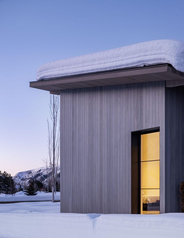 Exterior view of a compact modern building in a snowy alpine setting. The structure has vertically ribbed grey cladding, a broad flat projecting roof with deep eaves, and a tall black-framed glazed opening glowing warmly from the interior. Snow blankets the roof and ground, with bare trees and distant snow-covered mountains visible in the background.