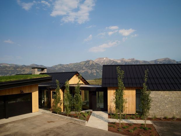 Exterior view of a contemporary mountain residence composed of several connected low-rise volumes. The building combines dark standing-seam pitched roofs, warm timber cladding, extensive glazing, and a pale natural-stone wall. Landscaped planting beds and paved forecourt sit in front, with rugged mountain ranges visible beyond.