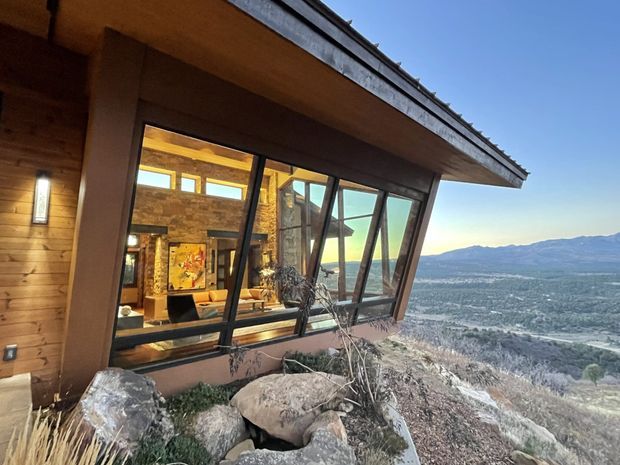 Exterior view of a contemporary mountain home set on a rocky, sloping site. The facade combines warm timber cladding with dark framed, angled floor-to-ceiling glazing beneath a projecting roof edge. Through the windows, a warm rustic living area with stone walls and timber finishes is visible, while a broad mountain valley landscape extends beyond the house.