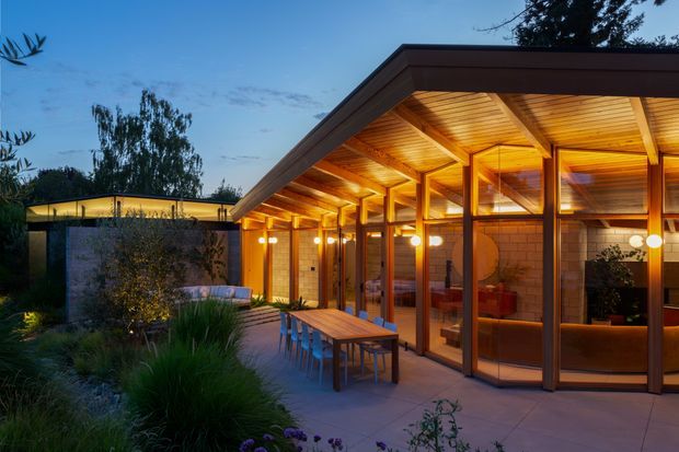 Dusk exterior view of a contemporary single-storey pavilion or extension with a broad low-pitched roof and extensive floor-to-ceiling glazing. Warm timber-lined soffits and exposed timber posts create a sheltered outdoor dining terrace, with a long timber table and chairs in front of an illuminated interior. Landscaped planting surrounds the paved terrace, and a separate flat-roofed structure is visible in the background.