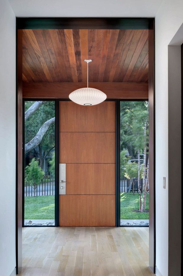 A contemporary residential entryway viewed from inside, featuring a tall pivot-style timber front door with matching narrow full-height sidelights, a richly stained timber-lined ceiling and upper wall surround, pale timber flooring, white walls, and a sculptural oval pendant light. The glazing opens toward a landscaped garden and mature trees outside.
