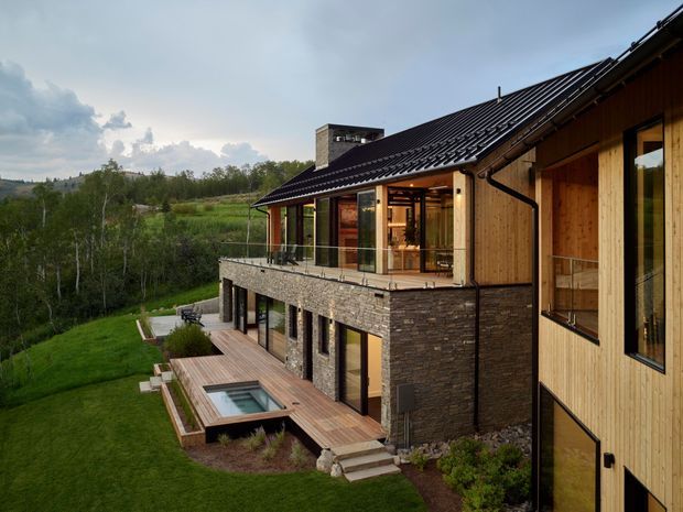 Exterior architectural view of a contemporary multi-level home set into a green, wooded landscape. The house combines warm vertical timber cladding, dark stone veneer, extensive glazing, a dark pitched metal roof, and a glass-balustraded upper terrace. A timber deck with a small rectangular plunge pool runs along the lower level.