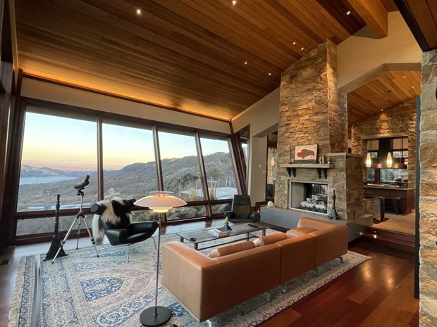 A spacious, warm living room with expansive angled floor-to-ceiling glazing overlooking a mountainous waterfront landscape. The room features a rich timber-lined ceiling with recessed spotlights, exposed beams, dark timber flooring, substantial natural-stone fireplace columns and surround, and a large cognac leather sectional arranged around a central coffee table. A patterned area rug, sculptural floor lamp, lounge chair, telescope, and layered fireplace joinery complete the refined lodge-like interior.