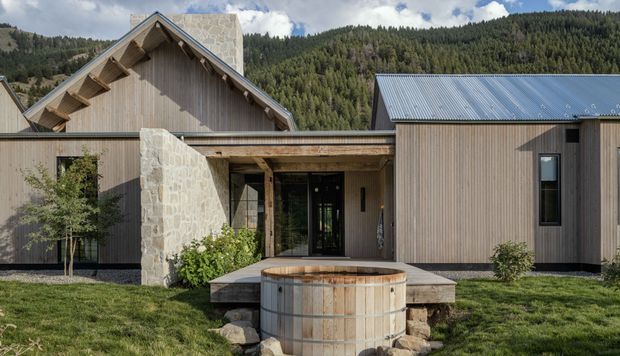 Exterior view of a contemporary mountain lodge or residential retreat composed of several connected gabled and low-pitched volumes. The facade combines vertical timber cladding, natural stone walls, dark-framed glazing, and corrugated metal roofing. A timber soaking tub sits in the foreground beside a covered entry, with forested mountains behind the building.