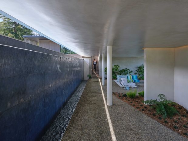 A covered exterior walkway runs alongside a dark textured boundary wall and landscaped garden beds. Slim white columns support a broad flat concrete roof, with a narrow strip of decorative stone at the wall base and a stairway leading upward in the distance. Outdoor seating is visible beneath the canopy, creating a sheltered transition space with strong linear perspective and contemporary detailing.