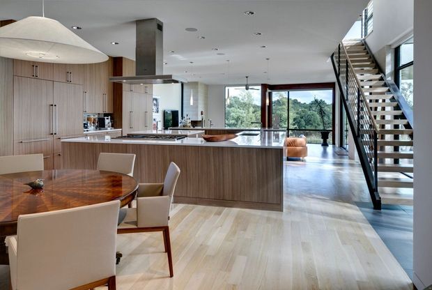 Open-plan contemporary kitchen and dining area with extensive light-wood cabinetry, a large central island with a pale stone benchtop, polished concrete flooring, and a stainless-steel rangehood. A round timber dining table occupies the foreground, while a black metal open-riser staircase leads to an upper level. Floor-to-ceiling glazing opens the space to leafy outdoor views, and a large sculptural pendant hangs above the dining area.