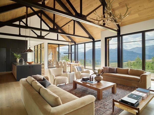 A spacious open-plan living room with a warm timber-lined ceiling, exposed dark timber or steel trusses, and expansive black-framed glazing overlooking a mountainous landscape. The room includes multiple cream and tan sofas and armchairs arranged around a substantial timber coffee table, with a dark kitchen island visible at the rear. A sculptural antler-style chandelier and pendant lights add rustic character to the refined contemporary interior.
