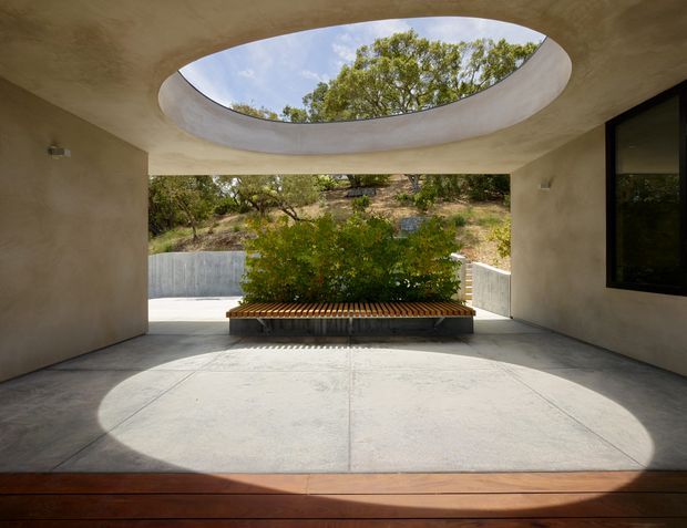 A sheltered, open-sided architectural courtyard or passage with smooth warm-toned plaster walls, a large oval skylight, concrete paving, and a built-in central planter bench. Sunlight creates a strong oval pool of light on the floor, while a dark-framed window is visible on the right and a landscaped hillside beyond the opening.
