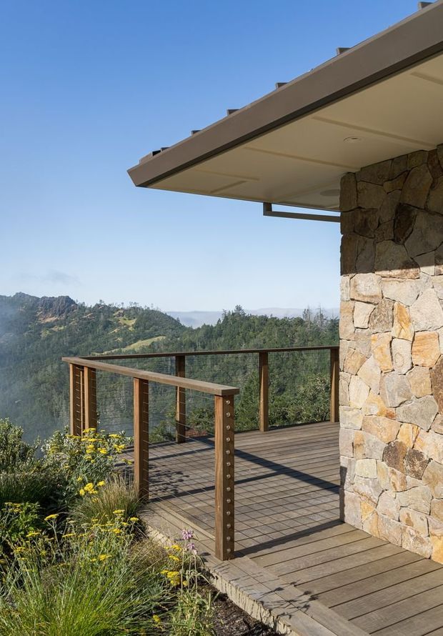 A contemporary mountain-view exterior terrace with dark timber decking, slim timber and metal balustrades, and a substantial irregular natural-stone wall supporting a broad low roof overhang. Native planting borders the deck, with wooded hills and open blue sky beyond. The image emphasizes the outdoor deck, rugged stone construction, and elevated scenic setting.