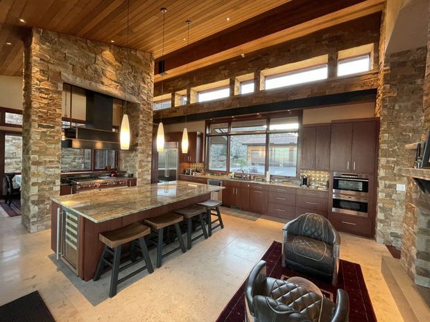 A spacious rustic-modern kitchen with a large stone-topped island and seating, dark timber cabinetry, exposed stone walls, substantial timber beams, and a warm timber slat ceiling. Pendant lights hang over the island, while a fireplace or cooking alcove is integrated into the stone wall. Leather lounge chairs and a rug occupy the foreground, and extensive glazing brings in natural light.