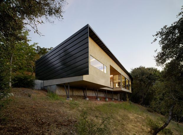Exterior view of a contemporary elevated house set within a wooded, sloping site. The building has a sharply pitched gable roof, dark horizontal cladding on the side elevation, a warm light-toned wall plane, extensive glazing, and a cantilevered upper volume supported by slender columns.