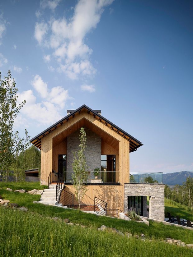 Exterior view of a contemporary alpine-style house set on a sloped, grassy site. The building has a prominent timber-clad gabled facade with deep exposed eaves, a central stone feature wall, extensive dark-framed glazing, and a lower-level stone volume with a terrace and glass balustrade. Mountainous terrain is visible in the distance.