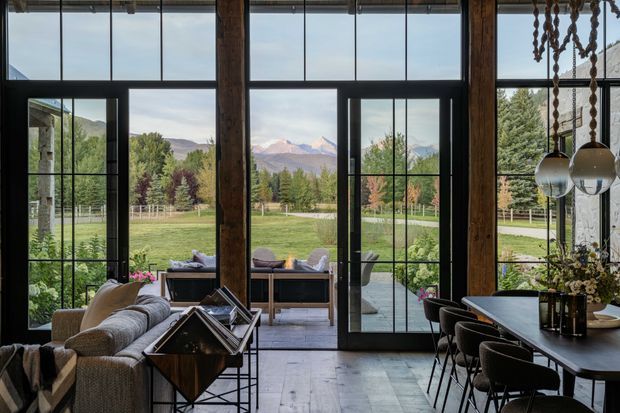 A rustic contemporary open-plan living and dining space with dramatic floor-to-ceiling black-framed glazing overlooking a landscaped rural valley and mountains. Exposed dark timber posts, large industrial-style windows and doors, a comfortable sectional sofa, pendant lighting and a long dining table create a warm, refined lodge-like interior with strong indoor-outdoor connection.