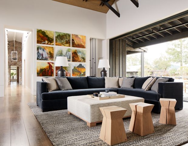 A spacious contemporary living room with a large dark navy sectional sofa, textured grey area rug, low upholstered rectangular coffee table, and sculptural light timber stools. White walls are decorated with a colourful grid of landscape artwork, while black-framed fully opening glazed doors connect the room to an outdoor area. Exposed dark roof beams and warm timber ceiling lining add architectural character.