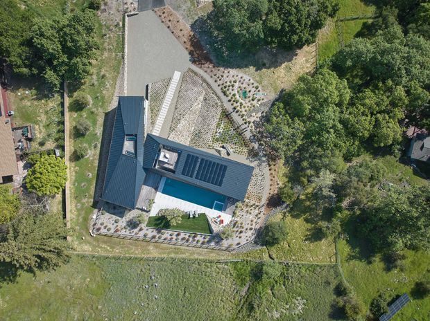 Aerial view of a dark-roofed residential property set within a landscaped, tree-lined site. The building has an irregular contemporary form, extensive dark metal roofing, a bright turquoise swimming pool, paved outdoor areas, and planted garden zones. Surrounding lawns, mature trees, and a small solar panel array are visible.