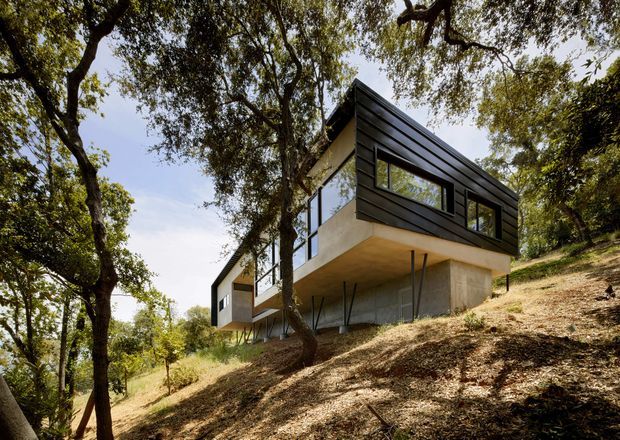 A contemporary elevated house set among mature trees on a steep, wooded hillside. The building has a cantilevered split-level form, dark horizontal cladding on the upper volume, pale concrete or rendered lower walls, large horizontal windows, and slender supports beneath the projecting structure.