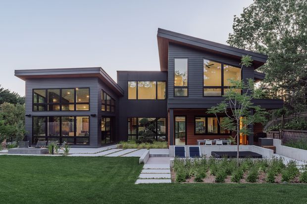 Exterior view of a contemporary multi-level residence with dark horizontal cladding, extensive floor-to-ceiling glazing, offset flat-to-low-pitched roof forms, and a landscaped front yard. The house includes projecting volumes, covered outdoor living areas, concrete paths, and large windows revealing warm-toned interiors.
