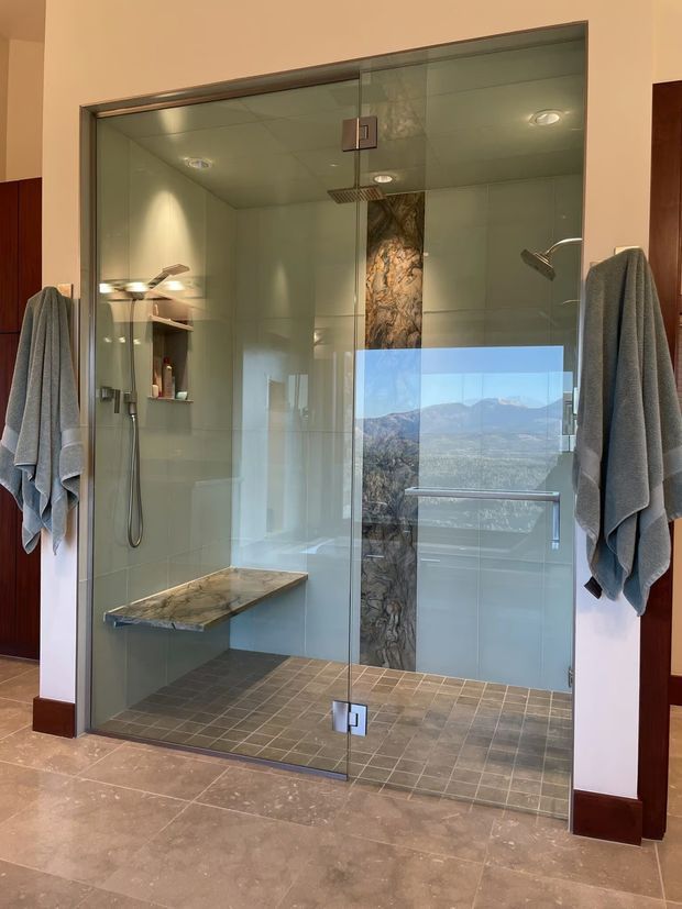 A spacious contemporary walk-in shower enclosure with frameless glass doors, chrome hardware, a wall-mounted rainfall shower and handheld shower, built-in shelving, and a floating stone bench. Large pale wall tiles contrast with smaller grey floor tiles, while a narrow marble-look feature strip runs vertically behind the shower. Grey towels hang on either side, and the glass reflects a mountainous outdoor view.