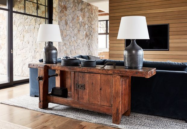 A warmly lit living room featuring a rustic, heavily timbered console behind a dark sofa. The room combines a pale irregular stone feature wall, horizontal timber wall panelling, black-framed glazing, timber flooring, and contemporary black table lamps. A television is mounted on the timber wall, with a woven neutral rug beneath the furniture.