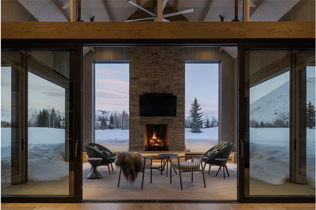 A warmly lit mountain-view living room centered on a stone-clad fireplace with a television above it. Large glazed sliding doors open toward a snowy outdoor landscape, while four dark lounge chairs and a low coffee table form a seating area around the fire. The room has exposed timber beams, a high pitched ceiling, and a mezzanine-like timber structure, creating a contemporary alpine retreat atmosphere.