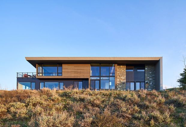 Exterior view of a contemporary two-storey residence set on a landscaped, sloping site. The house has a strong horizontal flat roof with deep eaves, extensive floor-to-ceiling glazing, dark metal framing, warm timber cladding, and contrasting natural stone feature walls. A small upper-level terrace is visible on the left, with dry native planting across the foreground.