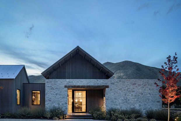 Exterior view of a contemporary mountain residence at dusk, composed of dark vertical timber-clad gabled wings and a central pale irregular-stone facade. Warm light glows through the entry glazing and side windows, with landscaped planting and a rugged mountain backdrop.