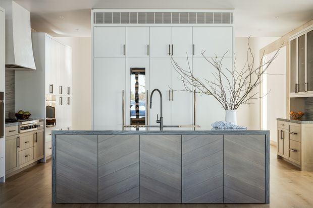 A bright, refined contemporary kitchen centred on a large island with diagonally grained grey timber cabinetry. The island includes an inset sink and dark gooseneck tap, while pale full-height cabinetry provides integrated storage and appliances behind it. Matching perimeter cabinetry, light benchtops, warm timber flooring, and understated hardware create a clean, tailored composition. Decorative branches in a vase add an organic accent.